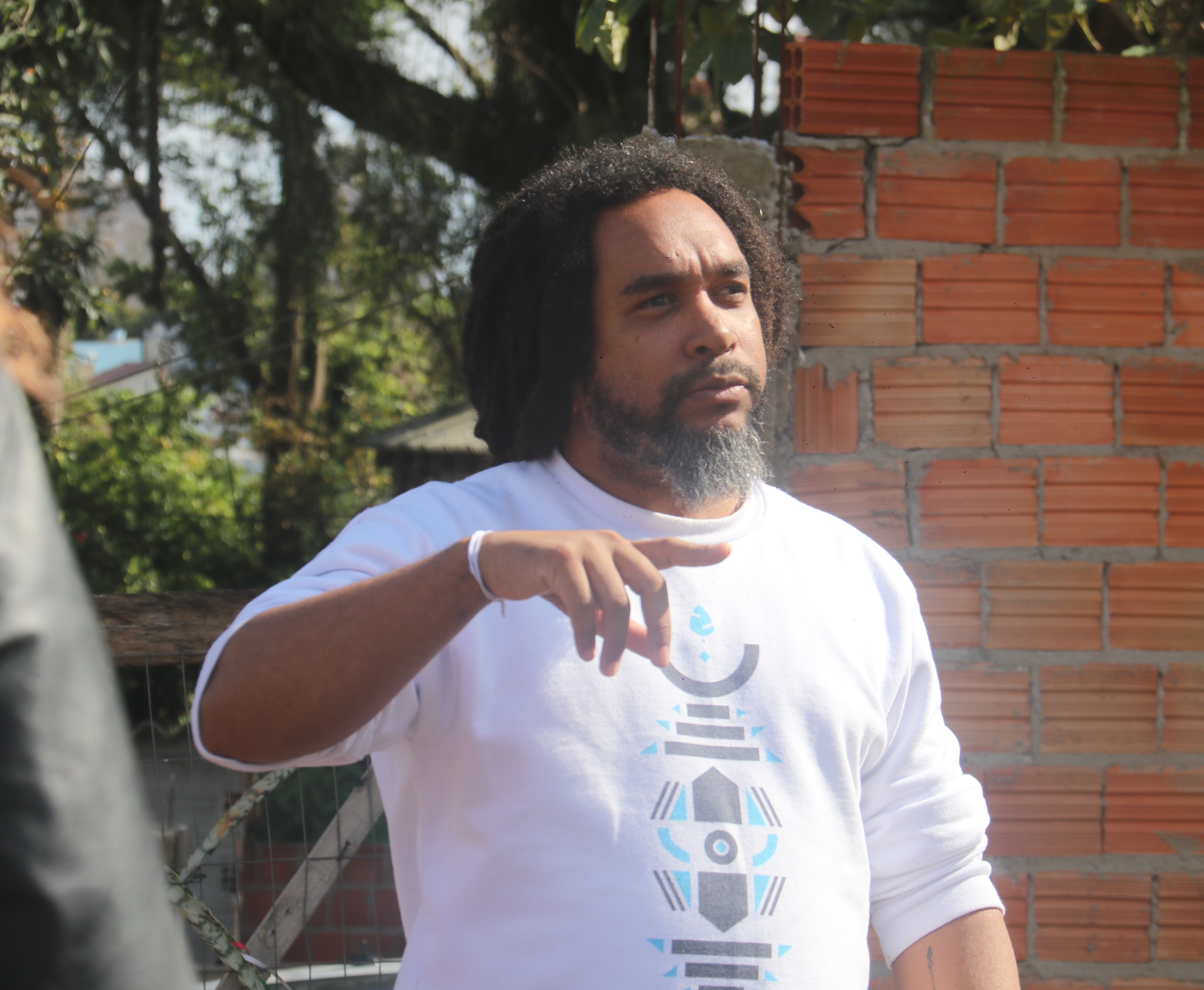 Imagem fotográfica em cores, na horizontal, mostra Gabriel Muniz, homem negro, com cabelo amarrado no estilo dreadlock, barba fechada preta, cavanhaque um pouco mais longo e grisalho. Ele está retratado em plano médio, em pé, vestindo um moletom branco com um desenho geométrico em tons de azul e preto. As mangas do moletom estão encolhidas de modo a mostrar os antebraços de Gabriel, e seu pulso direito é envolvido por uma fita de Senhor do Bonfim branca. O artista idealizador do projeto OuvidoChão está apontando com o referido braço para a direita, mesma direção para onde seu olhar é lançado. Ao fundo, há um muro de tijolos aparentes e algumas árvores. A composição é simples, focada na figura de Gabriel e no ambiente ao redor, que tem um ar natural e urbano ao mesmo tempo, o muro de entrada do Quilombo da Família Flores.