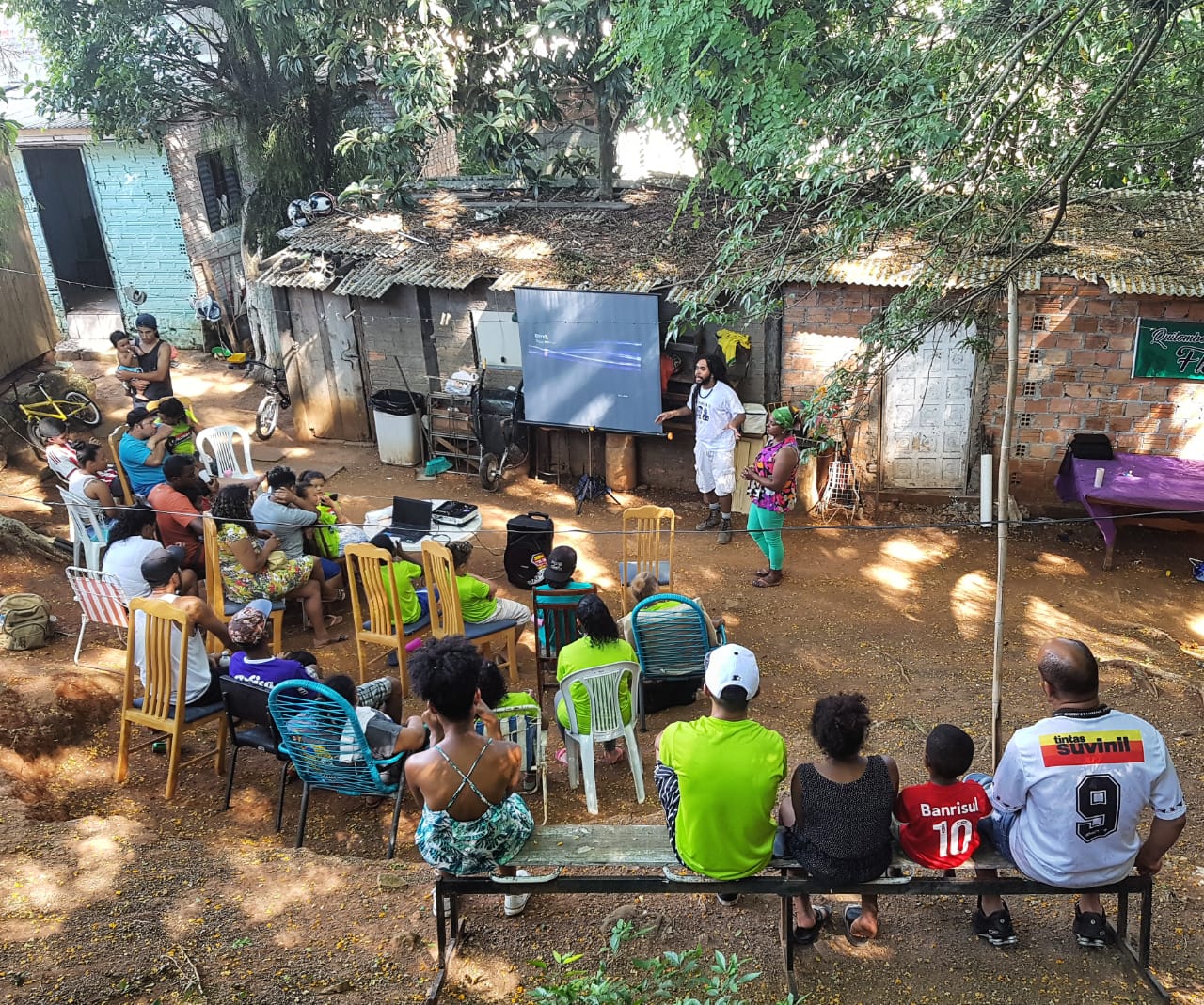 Fotografia colorida na horizontal, em plano geral, com iluminação natural do dia, de um grupo com cerca de 25 adultos e crianças de costas para nós, sentados em cadeiras. Vistos do alto, em um quintal de terra batida alaranjada, rodeado por árvores, observam Gabriel Muniz e Geneci Flores, que estão em pé à frente, ao lado de uma tela de projeção quadrada, montada em um tripé. À esquerda da tela, Gabriel gesticula enquanto fala. Veste camisa e bermuda de cor branca e calça uma bota marrom. Mais à esquerda, Geneci Flores em pé, usa um pequeno turbante verde amarrado na cabeça, veste uma blusa com estampa em tons de roxo e uma calça legging verde. Ao fundo, há duas casas de tijolos com telhas Brasilit.