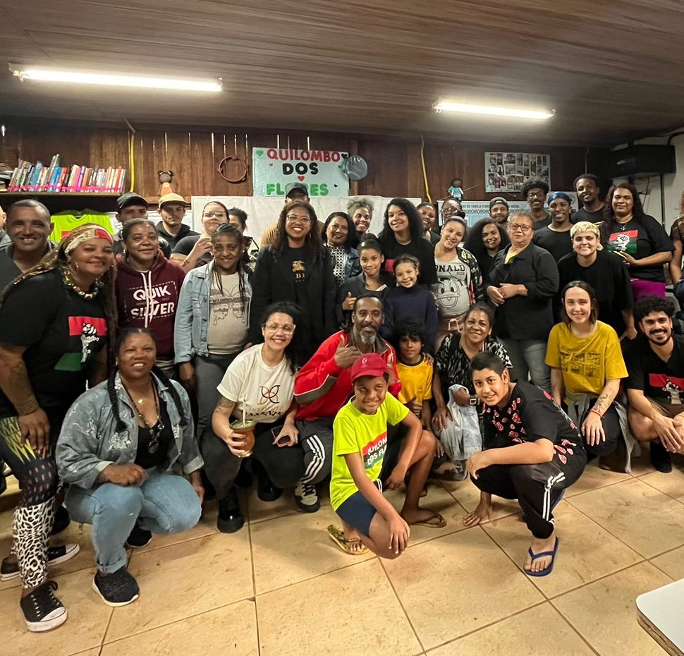 A fotografia em cores, na horizontal, mostra um grupo de familiares, moradores e apoiadores do Quilombo Flores. As pessoas estão reunidas em um ambiente fechado, um galpão com paredes de madeira que funciona como sede da associação comunitária. Há várias cadeiras e mesas dispostas ao redor. Ao fundo, uma estante com livros e um banner com a inscrição Quilombo dos Flores. As pessoas estão em pé, e algumas estão agachadas na frente, posando para a foto. Elas vestem roupas variadas e casuais. A iluminação é fornecida por lâmpadas fluorescentes no teto. A imagem destaca a união e colaboração entre as pessoas em um espaço coletivo, durante a finalização de uma das atividades organizadas pela comunidade.