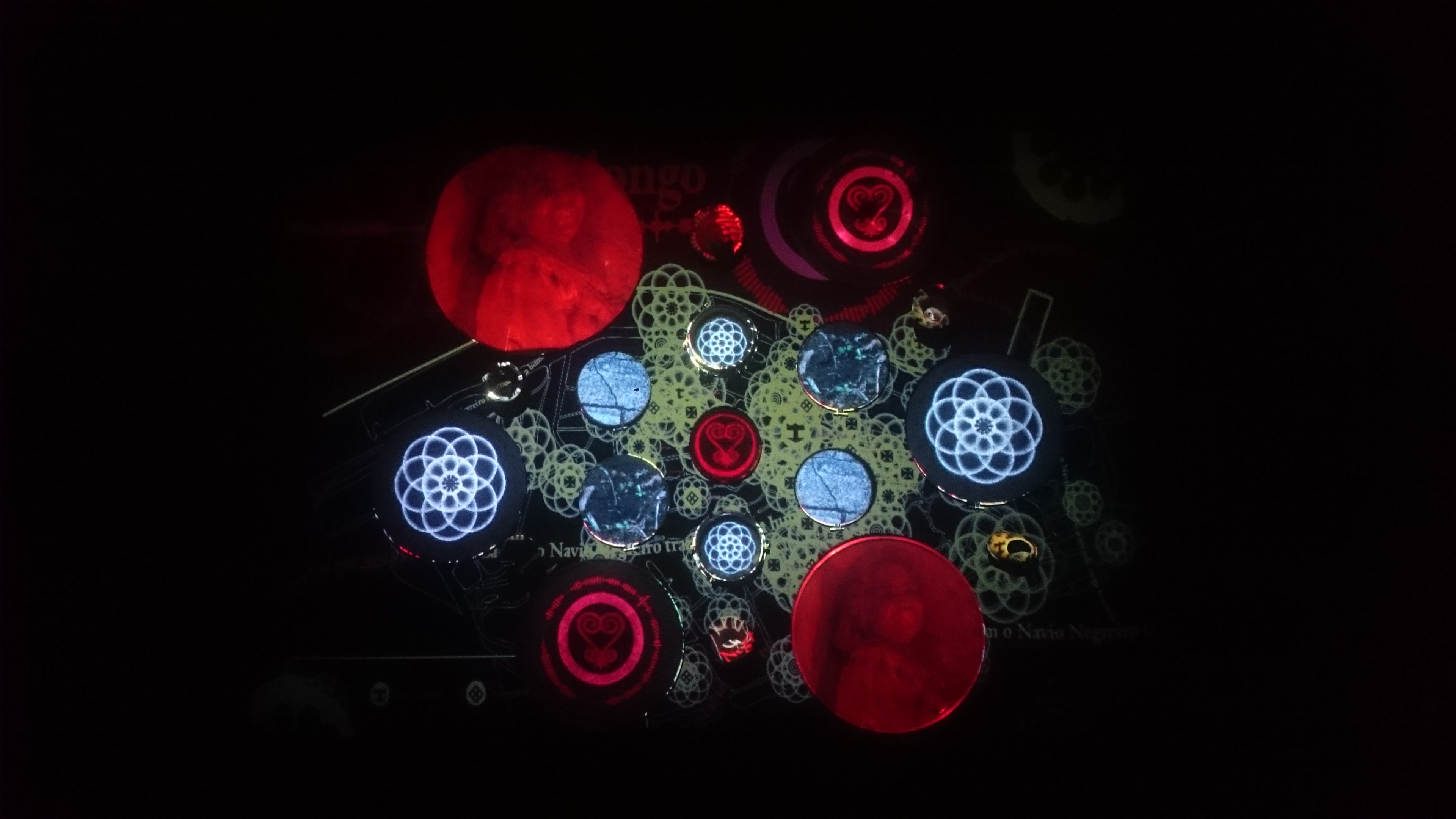 A fotografia em cores, na horizontal, mostra uma composição visual complexa e abstrata, com vários elementos circulares e padrões geométricos. Há círculos grandes e pequenos, todos eles contêm imagens ou símbolos projetados com videomapping. Dois dos círculos maiores, um no canto superior esquerdo e outro no canto inferior direito, são preenchidos com uma cor vermelha e contêm a imagem de uma mesma mulher negra. Outros círculos contêm padrões geométricos intrincados, como formas de flores ou mandalas – semente da vida – em cores como branco e azul. Há também círculos menores com símbolos adinkras. Entre eles, ao centro do painel, um círculo com o adinkra Sankofa. O fundo é escuro, o que faz com que os elementos coloridos se destaquem. A imagem é uma projeção de arte digital sobre um painel que compôs a instalação de arte multimídia OuvidoChão - Cartas Quilombolas, e apresenta uma complexidade visual pela combinação de elementos figurativos e abstratos, a saber: simbologias afrodiaspóricas, retrato de pessoas, elementos de cartografia e de geometria sagrada.