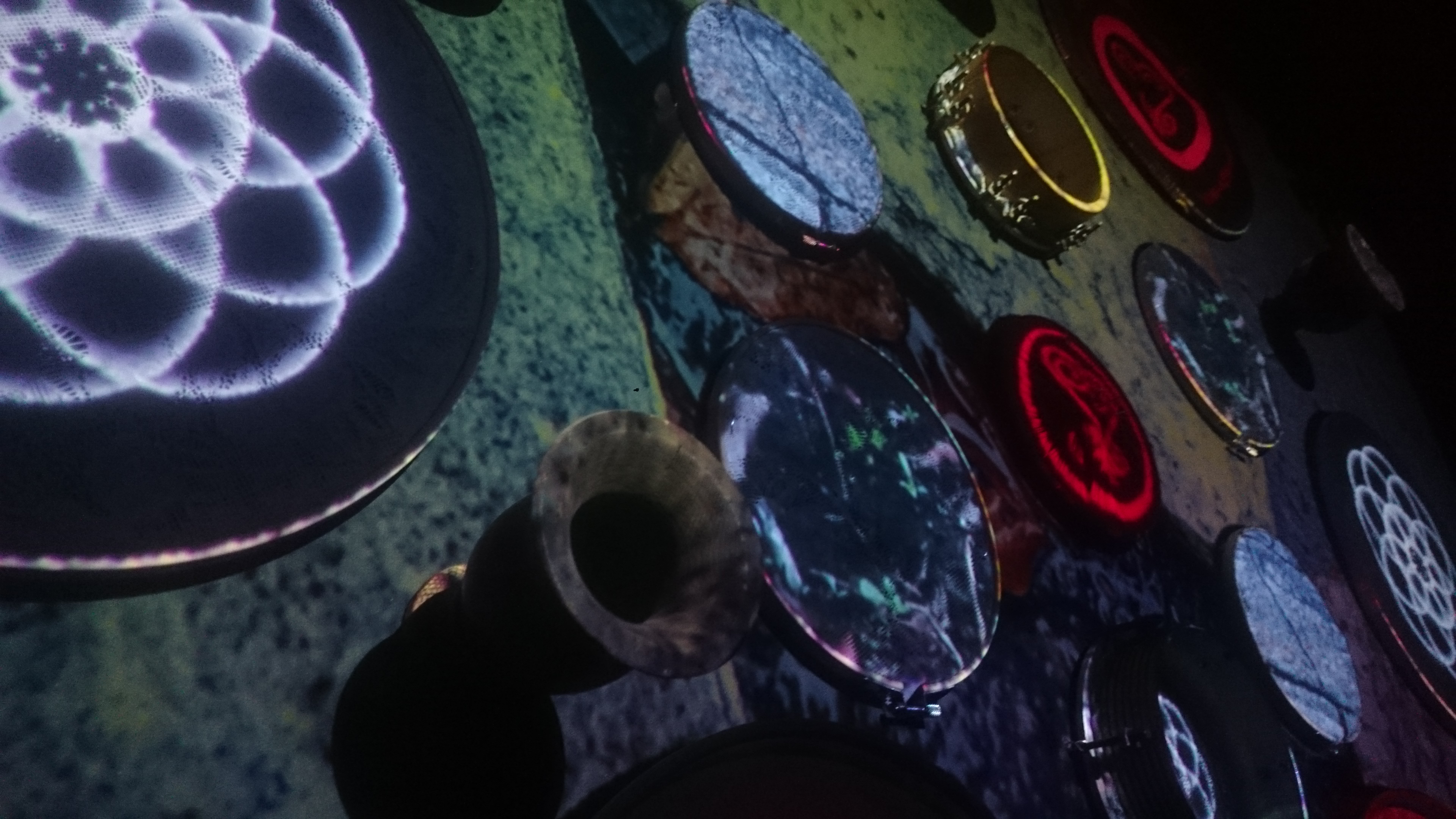 A imagem fotográfica em cores, em ângulo oblíquo de baixo para cima, mostra uma coleção de tambores, cuias de chimarrão e instrumentos de percussão dispostos em uma superfície, dentro de uma sala escura. Os tambores têm diferentes padrões e texturas projetadas com videomapping, incluindo formas geométricas e cores vibrantes, como vermelho, azul e branco. Alguns tambores têm padrões circulares complexos e símbolos adinkras, enquanto outros exibem texturas que lembram superfícies de pedra e um chão de bosque. A iluminação e as projeções criam um efeito visual interessante e dinâmico, destacando os detalhes dos instrumentos. A imagem representa a combinação de arte visual e sonora que compôs a instalação artística multimídia OuvidoChão - Cartas Quilombolas.