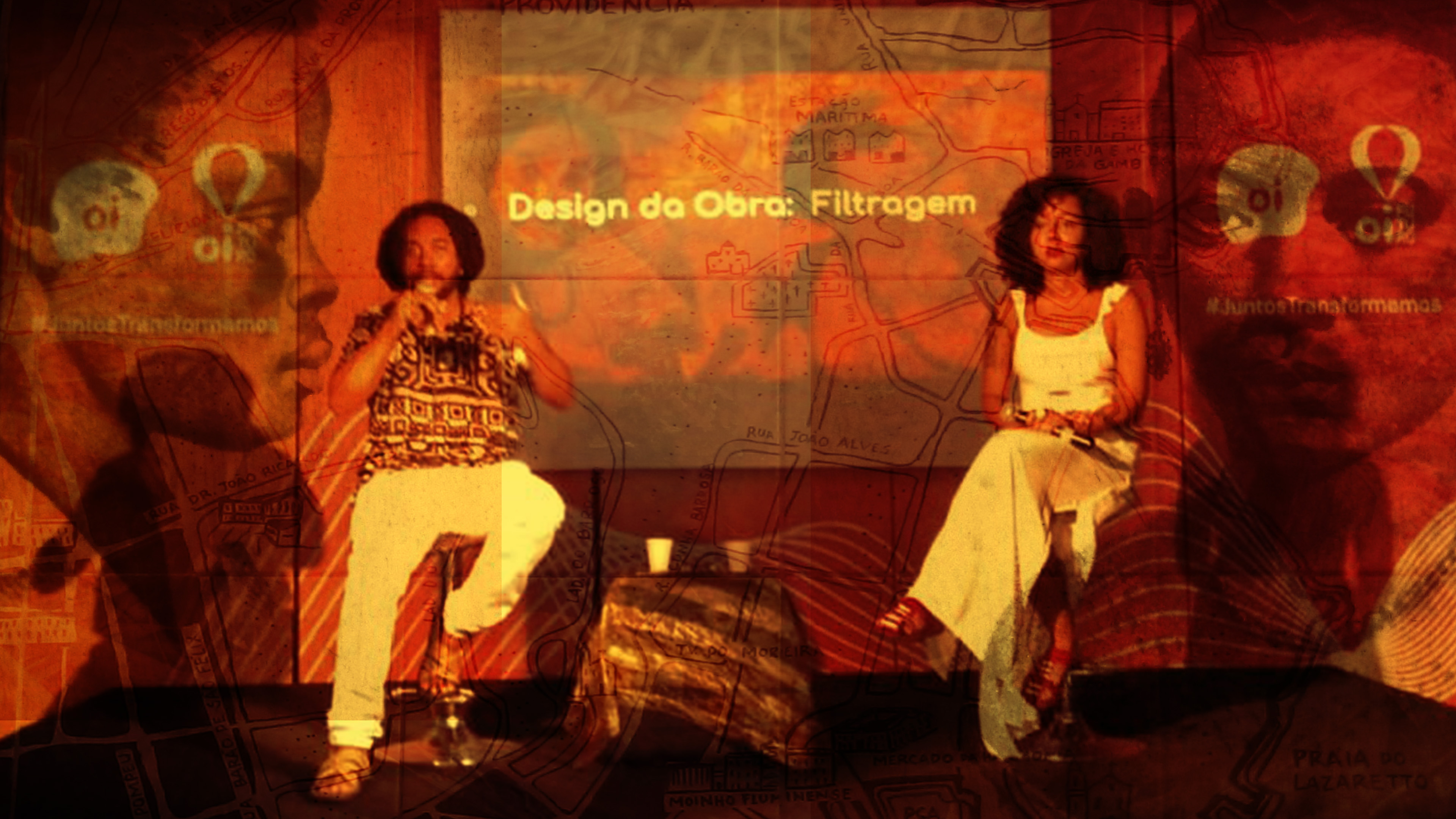 A fotografia em cores, na horizontal, mostra duas pessoas sentadas em cadeiras em um palco, participando de um evento sobre o projeto OuvidoChão - Cartas Quilombolas. À esquerda, sentado em uma cadeira alta, com a perna esquerda levemente dobrada, está Gabriel Muniz, homem negro, com cabelo amarrado, no estilo dreadlock. Veste uma bata estampada com motivos geométricos em tons de cinza, uma calça branca e alpercatas marrons. À direita, também em uma cadeira alta, com as pernas cruzadas, está Irla Franco, mulher negra, cabelos pretos encaracolados na altura do ombro. Ela veste um longo vestido branco sem mangas e calça uma sandália de tiras finas. No fundo, há uma projeção com o texto Design da Obra: Filtragem. O cenário parece ser uma combinação de elementos visuais, incluindo um mapa e imagens sobrepostas, criando um efeito artístico. Ambos estão segurando microfones, sugerindo que estão falando ou participando de uma discussão. A imagem combina elementos visuais e textuais de maneira criativa, para representar a dupla de artistas e para destacar um tema específico relacionado ao design e filtragem da obra em desenvolvimento.